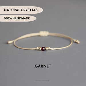 May include: A beige cord bracelet featuring a single dark red garnet bead flanked by two small gold-toned metallic beads. The text NATURAL CRYSTALS, 100% HANDMADE, and GARNET are visible. The bracelet has an adjustable sliding knot closure.
