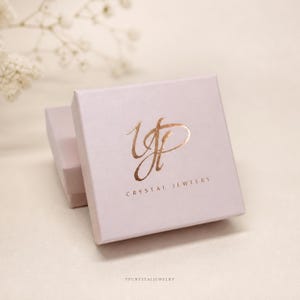 Puede incluir: Una caja de joyer&iacute;a cuadrada de color rosa claro con un logotipo de l&aacute;mina de oro rosa met&aacute;lico en la tapa. El logotipo muestra letras cursivas estilizadas y el texto CRYSTAL JEWELRY impreso en una fuente sans-serif debajo del dise&ntilde;o.