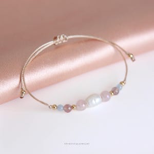 May include: A delicate adjustable bracelet featuring a central white oval pearl flanked by light pink, translucent rose-toned, and pale blue beads, accented with small gold-colored metal spheres on a thin beige cord. The text YPCRYSTALJEWELRY is visible.