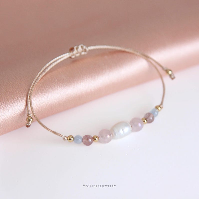 May include: A delicate adjustable bracelet featuring a central white oval pearl flanked by light pink, translucent rose-toned, and pale blue beads, accented with small gold-colored metal spheres on a thin beige cord. The text YPCRYSTALJEWELRY is visible.