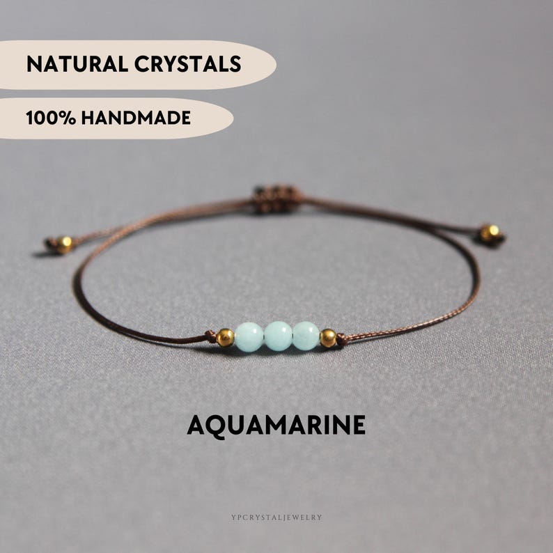 K&ouml;nnte beinhalten: Ein Armband aus brauner Kordel mit drei hellblauen Aquamarinperlen, die von zwei kleinen goldfarbenen Metallperlen flankiert werden. Das Armband hat einen verstellbaren Schiebeknoten und den Text NATURAL CRYSTALS, 100% HANDMADE und AQUAMARINE.