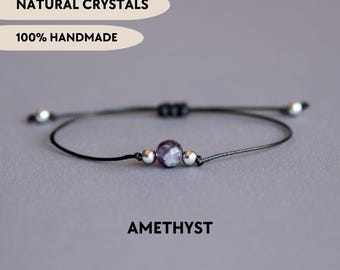 Puede incluir: Una pulsera de cordón negro con una cuenta de amatista morada facetada flanqueada por dos pequeñas cuentas metálicas plateadas. El texto NATURAL CRYSTALS, 100% HANDMADE y AMETHYST es visible. La pulsera tiene nudos deslizantes.
