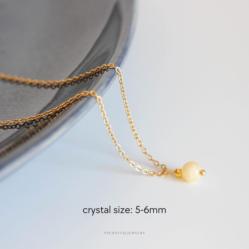 Puede incluir: Un collar de cadena en tono dorado con un colgante de una sola cuenta de cristal redonda de color amarillo p&aacute;lido. La cuenta est&aacute; acentuada por un peque&ntilde;o espaciador de metal dorado. El texto "crystal size: 5-6mm" y "ypcrystaljewelry" aparece debajo.
