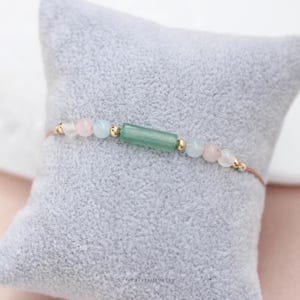May include: A delicate bracelet featuring a central green rectangular bead, flanked by pink, light blue, and clear round beads. Gold-colored accents and a thin, brown cord complete the design. The bracelet rests on a gray, textured surface.