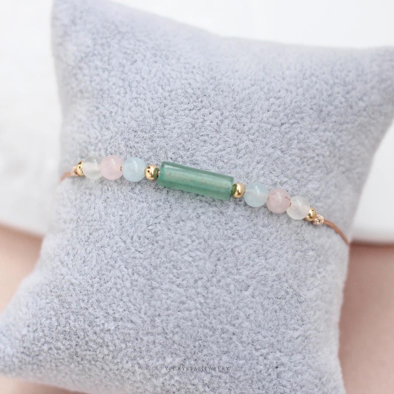 May include: A delicate bracelet featuring a central green rectangular bead, flanked by pink, light blue, and clear round beads. Gold-colored accents and a thin, brown cord complete the design. The bracelet rests on a gray, textured surface.