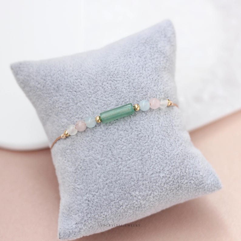 May include: Delicate bracelet featuring a central green rectangular bead, flanked by pink and blue round beads, and gold accents. The bracelet is strung on a thin, light-brown cord. The jewelry is displayed on a gray pillow.