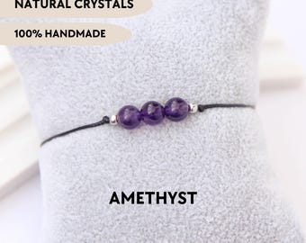 Puede incluir: Una pulsera con tres cuentas redondas de amatista morada separadas por pequeños espaciadores de metal plateado en un cordón negro fino. El texto NATURAL CRYSTALS, 100% HANDMADE y AMETHYST es visible. El diseño es sencillo y directo.