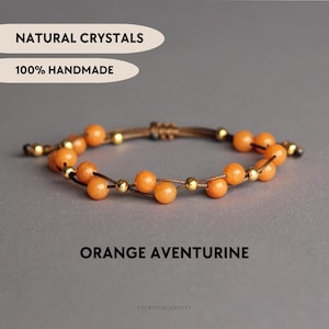 Puede incluir: Una pulsera con cuentas de aventurina naranja y pequeñas cuentas metálicas doradas en un cordón marrón. El texto NATURAL CRYSTALS, 100% HANDMADE y ORANGE AVENTURINE es visible. El diseño incluye un cierre de nudo corredizo ajustable.