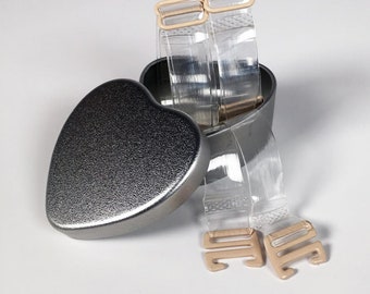 Puede incluir: Dos tirantes de sujetador de plástico transparente con cierres ajustables de color carne en una lata de corazón de plata. La lata tiene una superficie texturizada. El texto "ENDER LEGARD® WEAR YOUR FASHION WITH CONFIDENCE" está impreso en la parte inferior de la imagen.