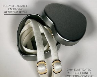 May include: Two white elastic bra straps with gold-colored metal hooks. The straps are packaged in a heart-shaped tin. The text on the tin reads "Fully recyclable packaging heart shape tin". The text on the straps reads "7mm elasticated and cushioned for extra comfort".