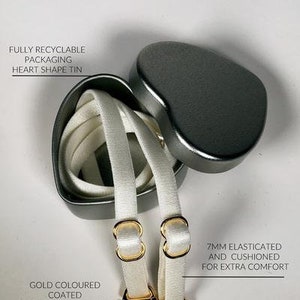 May include: Two white elastic bra straps with gold-colored metal hooks. The straps are packaged in a heart-shaped tin. The text on the tin reads "Fully recyclable packaging heart shape tin". The text on the straps reads "7mm elasticated and cushioned for extra comfort".