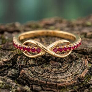 Op de afbeelding: Een goudkleurige infinity ring met een rij kleine, rode edelstenen. De ring is geplaatst tegen een getextureerde, bruine achtergrond, met een vage groene achtergrond.