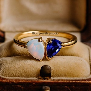 Könnte beinhalten: Ein goldener Ring mit zwei tropfenförmigen Edelsteinen: einem weißen Opal und einem blauen Saphir. Der Ring wird in einer Vintage-Schmuckschatulle aus braunem Leder mit Samtinnenfutter präsentiert. Der Ring ist von Blushjewelrystudio.