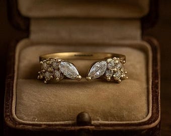 May include: A gold ring with a split shank design, featuring two marquise-cut stones and smaller accent stones. The ring is displayed in a vintage brown jewelry box with a velvet interior, set against a dark background.