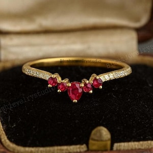 May include: A gold ring with a curved design, set with small red gemstones and tiny clear stones. The ring is displayed in a brown jewellery box with a black interior. The ring's design features a central heart-shaped ruby.