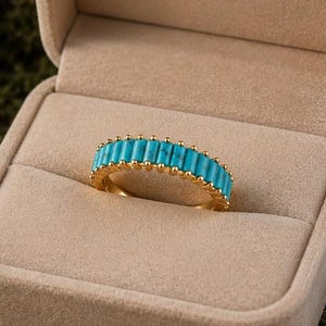 Könnte beinhalten: Ein goldener Ring mit einem Band aus rechteckigen türkisfarbenen Steinen wird in einer beigen Ringschatulle präsentiert. Die Schatulle ist geöffnet und zeigt den Ring vor einem weichen, samtigen Innenraum. Der Text "BlushJewelryStudio" ist auf der Innenseite der Schatulle aufgedruckt.