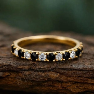 Peut inclure: Une bague en or ornée d'une rangée alternée de pierres noires et blanches. La bague est posée sur une surface en bois, mettant en valeur le contraste entre l'or et les pierres. Un bijou élégant pour toutes les occasions.