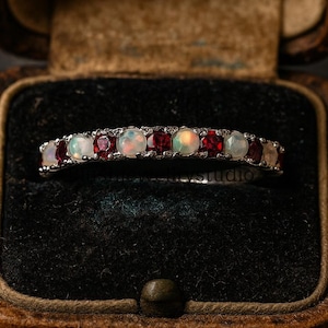 Puede incluir: Un anillo de plata con ópalos y rubíes alternados, presentado en un joyero vintage de cuero marrón con interior oscuro. El anillo es una banda delicada con pequeñas piedras redondas.