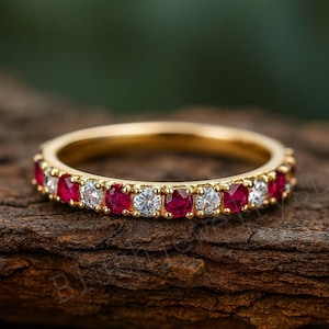 May include: A gold band ring with alternating red and clear gemstones. The ring is set on a textured, brown surface. The gemstones are evenly spaced around the band, creating a continuous pattern. The ring is a piece of jewelry.