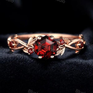 May include: A rose gold ring with a red gemstone and leaf accents. The ring is set on a black background.