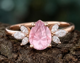 May include: A rose gold ring featuring a pear-shaped pink gemstone, flanked by six marquise-cut white gemstones. The ring is set against a textured, brown surface, with a blurred green background.