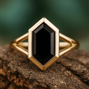 May include: A gold ring with a black, hexagonal gemstone. The ring's band has a split design on each side of the gemstone. The ring is set on a piece of textured wood, with a blurred green background.