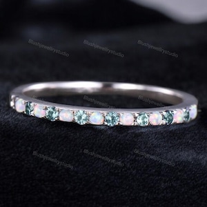 May include: A silver ring with a band of alternating white and teal gemstones. The gemstones are round and evenly spaced around the ring. The ring is set against a dark background.