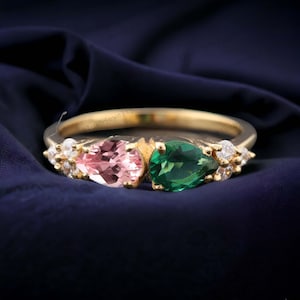 Könnte beinhalten: Ein goldener Ring mit zwei birnenförmigen Edelsteinen, einem rosa und einem grünen, umgeben von kleineren weißen Edelsteinen.