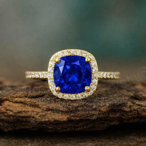 May include: A gold ring with a large, square, deep blue gemstone, encircled by smaller, clear stones. The ring's band is also set with small, clear stones. The ring is displayed on a textured, brown surface.
