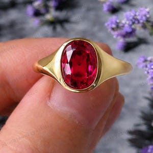 May include: A gold ring featuring a large, oval-cut ruby gemstone. The ruby is a vibrant red color and is set in a polished gold band. The ring's design is simple yet elegant, showcasing the beauty of the gemstone.