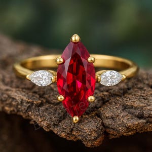 Puede incluir: Un anillo de oro con una gran gema roja intensa en forma de marquesa como pieza central. El anillo está adornado con piedras blancas más pequeñas en forma de pera a cada lado de la piedra central. El anillo está sobre una superficie oscura y texturizada.