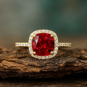 May include: A gold ring with a large, square-cut, vibrant red gemstone surrounded by a halo of smaller, clear stones. The band is also adorned with small, clear stones. The ring is set against a textured, brown surface.