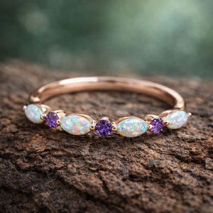 Puede incluir: Anillo de oro rosa con ópalos ovalados y piedras púrpuras redondas alternados. Los ópalos tienen un aspecto blanco lechoso e iridiscente, mientras que las piedras púrpuras son de un violeta intenso. El anillo está sobre una superficie de madera texturizada.