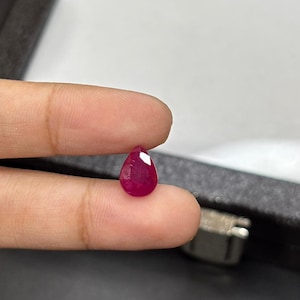 Pode incluir: Uma gema de rubi facetada em forma de gota. A gema é de uma cor vermelha profunda e saturada, com uma superfície polida que reflete a luz. O rubi é segurado entre dois dedos, mostrando seu tamanho e forma.