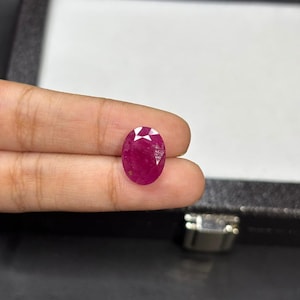 May include: A faceted, oval-shaped ruby gemstone. The ruby is a deep, vibrant red color with visible internal inclusions. The gemstone is held between fingers against a dark background.