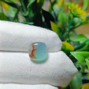 May include: A small, square-shaped gemstone with a light blue hue and reddish-brown inclusions. The stone is held against a white fabric background, with green and yellow foliage visible in the background.