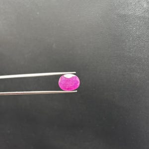 May include: A vibrant, oval-cut pinkish-red gemstone held by a pair of silver-toned metal tweezers. The faceted stone displays a brilliant, translucent quality with visible internal inclusions and a polished surface reflecting light.