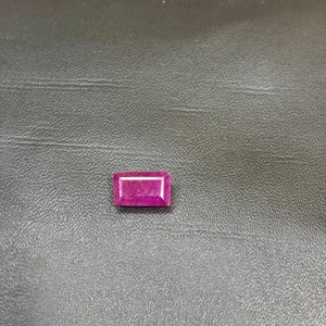 May include: A rectangular cut gemstone with a vibrant magenta-pink hue. The stone features a faceted surface that reflects light, showcasing a clear, polished appearance with sharp, defined edges and a symmetrical geometric shape.