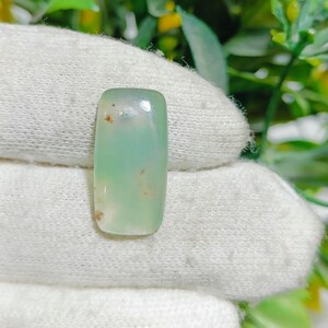 May include: A rectangular, polished gemstone with rounded edges. The stone has a translucent, pale green hue with subtle brown inclusions. The gemstone is set against a white, textured background, with green foliage visible in the background.