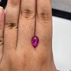 May include: A vibrant, oval-shaped, cabochon-cut gemstone with a deep magenta-pink hue. The stone features a smooth, polished, domed surface that reflects light, showcasing a translucent quality and internal inclusions within the crystal structure.