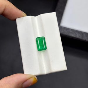 以下が含まれることがあります： 鮮やかな緑色の長方形のエメラルド。宝石は白い背景に配置されています。エメラルドは手で持たれており、その透明度と輝きを際立たせています。この宝石は貴重な宝石です。