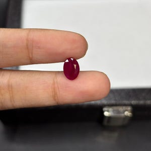 May include: A polished, oval-shaped ruby gemstone. The deep red gem is held between fingers, highlighting its facets and rich colour. The ruby is a symbol of passion and luxury.