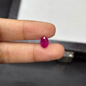 以下が含まれることがあります： ファセット加工された、楕円形のルビーの宝石。宝石は鮮やかで深い赤色で、2本の指の間に挟まれています。ルビーは研磨された表面を持ち、光を反射するファセットが見えます。背景は暗く、ニュートラルな色調です。