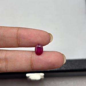May include: A loose, oval-cut ruby gemstone with a deep, saturated red colour and a faceted surface that reflects light. The gemstone is small and displays a clear, transparent quality with a polished finish, showcasing its natural brilliance.