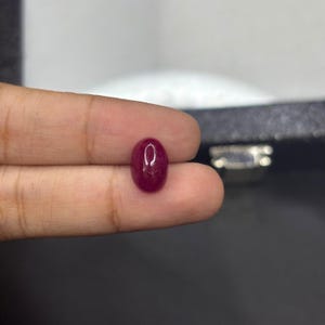 Peut inclure: Une pierre précieuse de forme ovale, un rubis poli. Le rubis est d'un rouge profond avec une surface lisse et réfléchissante. La gemme est tenue entre les doigts, mettant en valeur sa taille et sa forme. Idéal pour la création de bijoux.