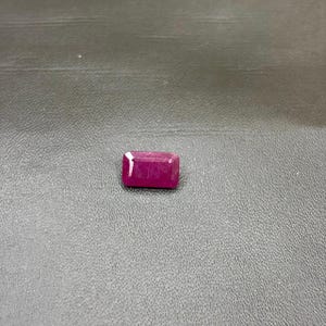 Puede incluir: Una piedra preciosa rectangular facetada de un color rojo magenta profundo y vibrante. La piedra presenta un diseño clásico de talla esmeralda con bordes limpios y afilados, y una superficie pulida y reflectante que capta la luz en su parte superior.