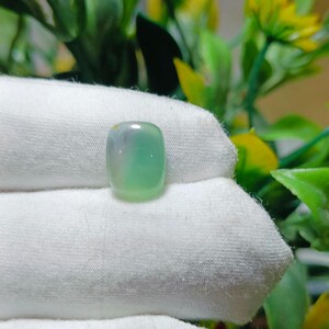 Peut inclure: Une pierre précieuse de jadéite vert clair translucide de forme rectangulaire arrondie. La pierre a une surface lisse et polie et un léger dégradé de couleur, passant d'un lavande pâle à un vert vif. La jadéite est présentée sur un tissu blanc.