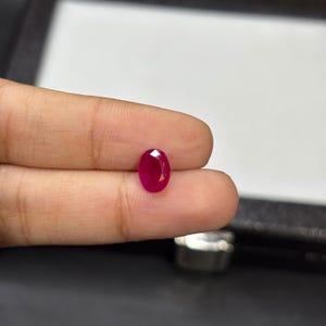 Puede incluir: Una gema de rubí facetada de forma ovalada. El rubí es de un color rojo intenso y vibrante y se sostiene entre los dedos. La gema se muestra sobre un fondo neutro, resaltando su brillo y claridad. Adecuado para la fabricación de joyas.
