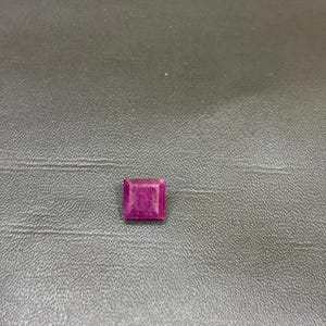 May include: A square-cut, deep magenta-colored gemstone with a faceted surface. The stone features a distinct four-sided shape with sharp corners and a polished finish, displaying internal light reflections across its vibrant, saturated pinkish-red surface.