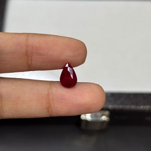 Puede incluir: Una gema de rubí facetada en forma de lágrima. La gema de color rojo intenso se sostiene entre dos dedos, mostrando sus superficies pulidas y su color brillante. La forma y el color del rubí sugieren que podría usarse en joyería.